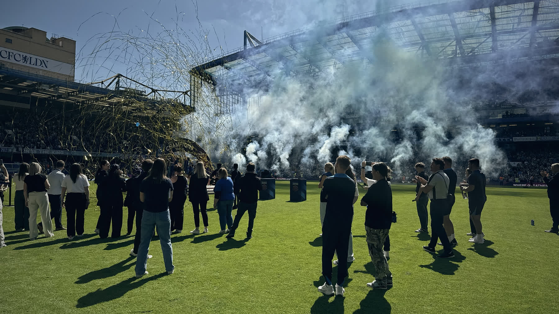 Gold streamers and blue smoke over the pitch at a trophy presentation in a full stadium.